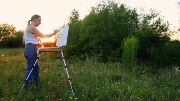 Una artista con un pincel y un polytray en sus manos dibuja sobre lienzo sobre el telón de fondo de la naturaleza. El artista se basa en el caballete. Artista en el trabajo — Vídeos de Stock