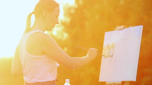 Ritratto di una ragazza con i capelli bianchi in una t-shirt bianca, raffigurante un paesaggio su tela al tramonto al sole con pittura ad olio e pennello . — Video Stock