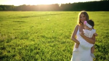 Anne ile kızı parkın içinden beyaz elbise ve kameraya bakarak gün batımında yürür.