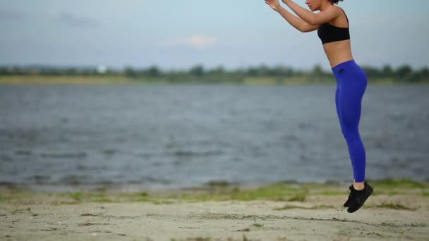 Bela menina de esportes faz burpees pulando na praia perto do rio fluindo . — Vídeo de Stock