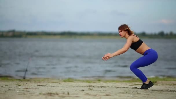 Bella ragazza sportiva fa burpees saltando sulla spiaggia vicino al fiume che scorre . — Video Stock