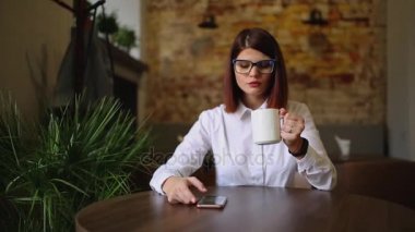 Simpatic esmer gözlük yakın çekim ile. İçecekler hoş kokulu kahve ve kullanarak cep telefonu metin iletileri çevirir, haberleri izliyor, fotoğraflar arasında çevirir, bir sosyal ağ iletişim kurar.