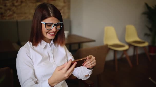 Glückliche junge Frau sitzt im Coffeeshop und schreibt SMS auf ihrem Smartphone — Stockvideo