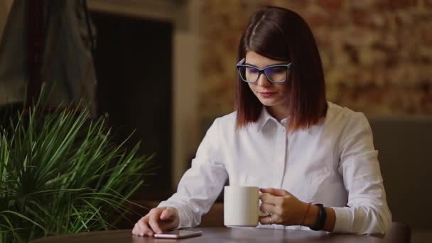 Simpatic brunette met bril close-up. Dranken geurende koffie en het gebruik van een mobiele telefoon belt tekstberichten horloges nieuws, gespiegeld door foto's, communiceert in een sociaal netwerk. — Stockvideo