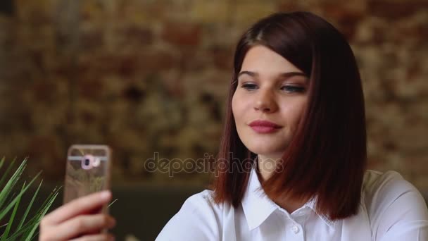 Een mooi meisje zit in een café en na een hete koffie, dronken vormt voor de camera van een mobiele telefoon te maken een mooi zelf voor sociale netwerken. — Stockvideo