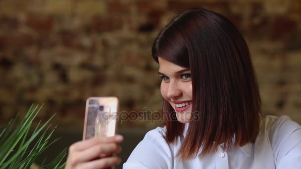 Une belle fille s'assoit dans un café et après avoir bu un café chaud, pose pour une caméra de téléphone portable pour faire un beau soi pour les réseaux sociaux . — Video