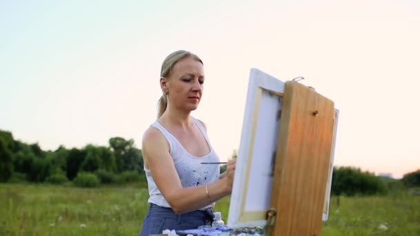 Een jonge vrouw met lang haar, dat zich in de wind ontwikkelt schetst een beeld op doek, dat op de ezel staat. De dame is in een open lucht, ze tekent uit het leven — Stockvideo