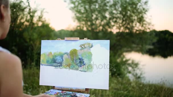 Jonge kunstenaar schilderij beeld in park met borstels en kleuren zitten door de romantische lake en stedelijke gebouwen in de achtergrond. ultra brede hoekmening — Stockvideo