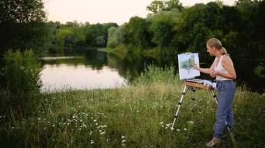 genç sanatçı Resim Resim fırçaları ve romantik göl ve arka plan kentsel binalarda oturan renkleri ile parkta. Ultra geniş açılı görünüş
