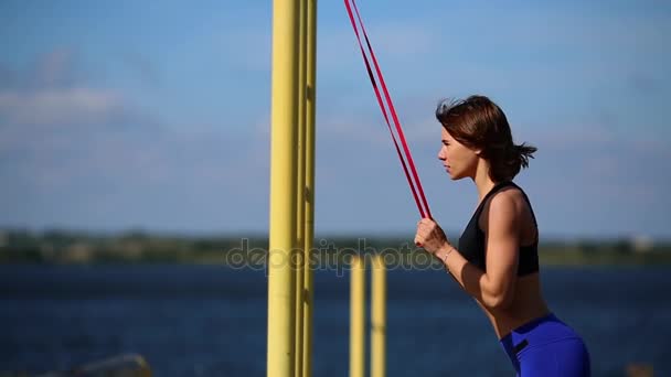 Bruna in leggings blu sulla riva del fiume esegue esercizi sui tricipiti con un elastico . — Video Stock