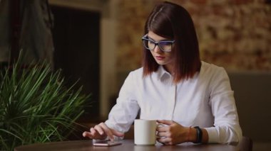 Simpatic esmer gözlük yakın çekim ile. İçecekler hoş kokulu kahve ve kullanarak cep telefonu metin iletileri çevirir, haberleri izliyor, fotoğraflar arasında çevirir, bir sosyal ağ iletişim kurar.