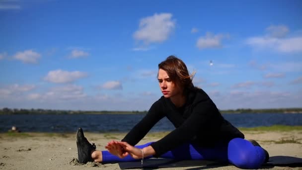 Fille sur la rive de la rivière effectue l'étirement et la relaxation des muscles après un entraînement dur. Fitness et yoga à proximité de la plage . — Video