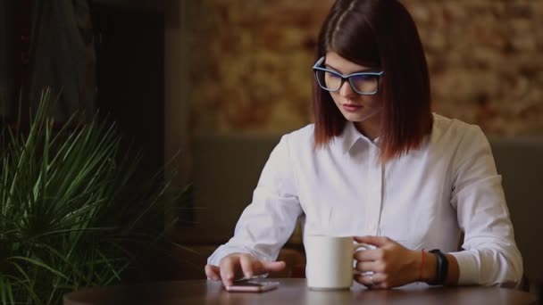 Simpatic brunette met bril close-up. Dranken geurende koffie en het gebruik van een mobiele telefoon belt tekstberichten horloges nieuws, gespiegeld door foto's, communiceert in een sociaal netwerk. — Stockvideo