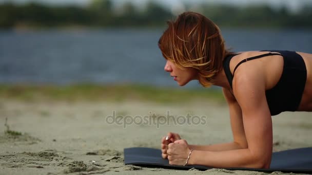 Красивая спортивная девушка делает упражнение на доске на берегу синего озера. Крупный план — стоковое видео