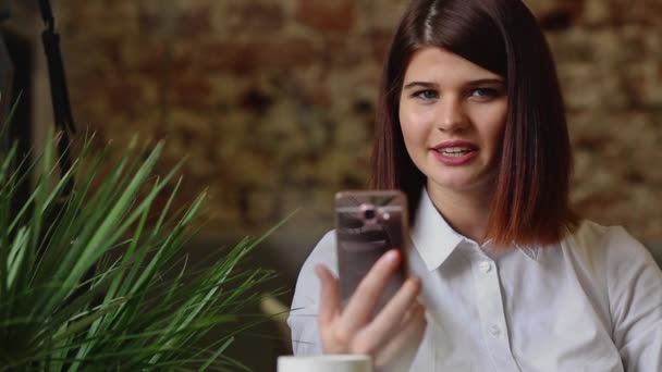 Sonriente chica en el café hablando en el chat de vídeo en el teléfono inteligente mientras bebe café — Vídeos de Stock