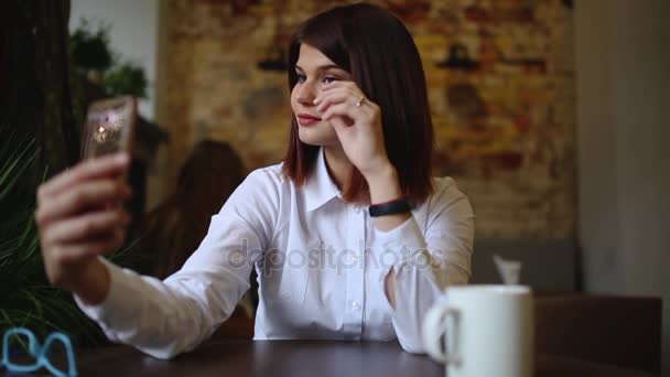 Uma linda garota se senta em um café e depois de beber um café quente, posa para uma câmera de telefone celular para fazer um belo self para redes sociais . — Vídeo de Stock