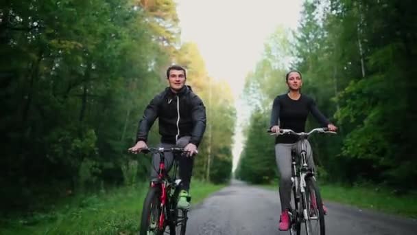 Handsome gebaarde mannelijke wielrenner zijn race-fiets in de ochtend samen met zijn vriendin, beide dragen beschermende helmen en brillen, de zon schijnt door ertussen. — Stockvideo