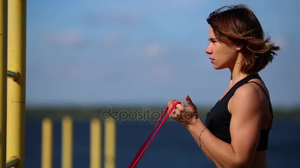 Morena desportiva bonito com leggings azuis realizar exercícios para fortalecer os músculos das mãos na rua com elástico elástico esticável. Exercício ao ar livre. Close-up — Vídeo de Stock