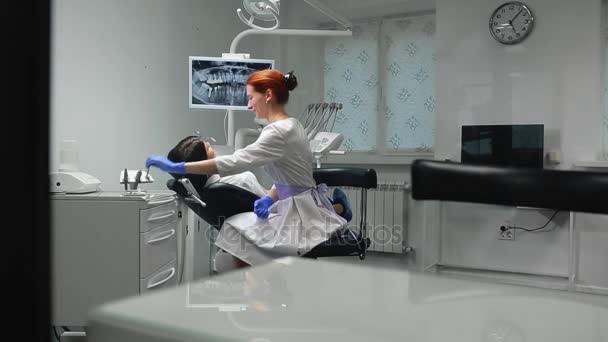 Female doctor in blue gloves and a white coat approaches to the chair of the patient and using x-ray and instruments the dentist inspects the teeth in the oral cavity. Girl patient. — Stock Video