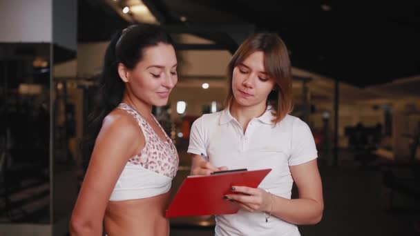 Entrenadora personal femenina hablando con una cliente en un gimnasio usando una tableta para notas . — Vídeos de Stock