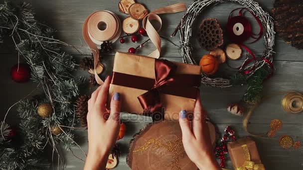 A top-down plan. Fully visible the table with the decorations. Female hands put and finalize Christmas gift wrapped in craftool paper on a wooden table. Bandaging tape and tied in a bow. — Stock Video