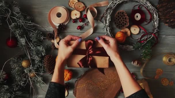 A top-down plan. Fully visible the table with the decorations. Female hands put and finalize Christmas gift wrapped in craftool paper on a wooden table. Bandaging tape and tied in a bow. — Stock Video