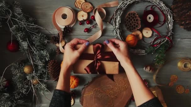 Un plan de arriba hacia abajo. Totalmente visible la mesa con las decoraciones. Las manos femeninas ponen y finalizan el regalo de Navidad envuelto en papel artesanal sobre una mesa de madera. Bandaging cinta y atado en un lazo . — Vídeo de stock