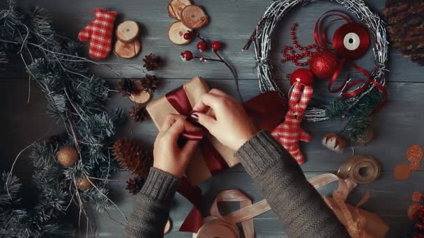 Um plano de cima para baixo. Totalmente visível a mesa com as decorações. As mãos femininas põem e finalizam o presente de Natal envolvido no papel craftool em uma mesa de madeira. Bandaging fita e amarrado em um arco . — Vídeo de Stock