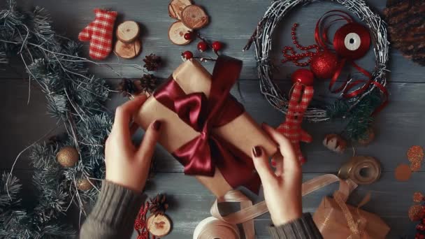 Un plan descendant. Entièrement visible la table avec les décorations. Les mains féminines mettent et finalisent le cadeau de Noël enveloppé dans du papier artisanal sur une table en bois. Bande de bandage et attaché dans un arc . — Video