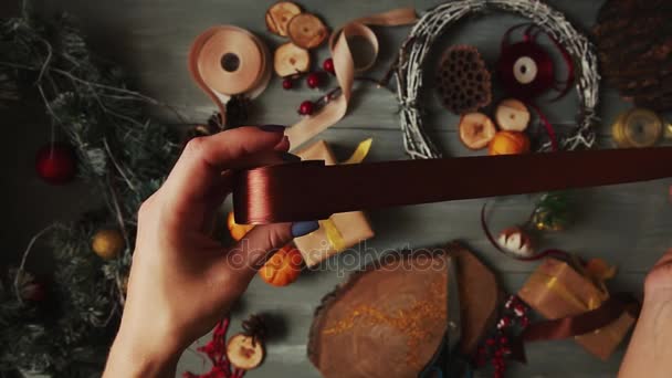 Een top-down-plan. Volledig zichtbaar in de tabel met de decoraties. Vrouwelijke handen zetten en finaliseren kerst cadeau verpakt in craftool papier op een houten tafel. Verbanden van tape en gebonden in een boog. — Stockvideo