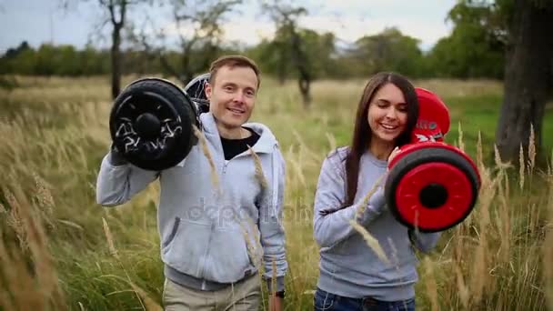 Young man and girl go into the field with hoverboards look at one another in urban clothing. Steadicam — Stock Video