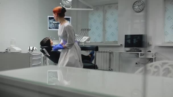 La doctora en guantes azules y una bata blanca se acerca a la silla del paciente y usando rayos X e instrumentos el dentista inspecciona los dientes en la cavidad oral. Chica paciente . — Vídeo de stock