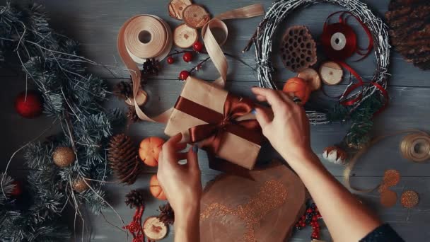 En uppifrån och ned-plan. Fullt synlig tabellen med dekorationer. Kvinnliga händer sätta och slutföra jul gåva insvept i craftool papper på ett träbord. Bandagen tape och knuten i en rosett. — Stockvideo