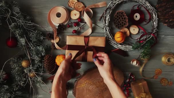 Un plan de arriba hacia abajo. Totalmente visible la mesa con las decoraciones. Las manos femeninas ponen y finalizan el regalo de Navidad envuelto en papel artesanal sobre una mesa de madera. Bandaging cinta y atado en un lazo . — Vídeo de stock