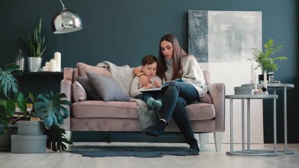 Mooie jonge moeder zittend op de Bank in uw huis de woonkamer leest zoon een verhaal in het oor en leren lezen van de kleine jongen. — Stockvideo