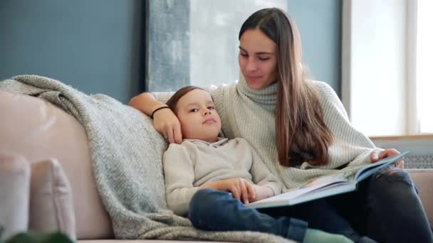 Mooie jonge moeder zittend op de Bank in uw huis de woonkamer leest zoon een verhaal in het oor en leren lezen van de kleine jongen. — Stockvideo