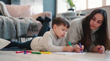 Oğlumla işaretleri ile ailesi tasvir kağıt üzerine çizim yerde yatarken bir sıcak kazaklı güzel genç anne. Çocuk öğrenir çizmek için.
