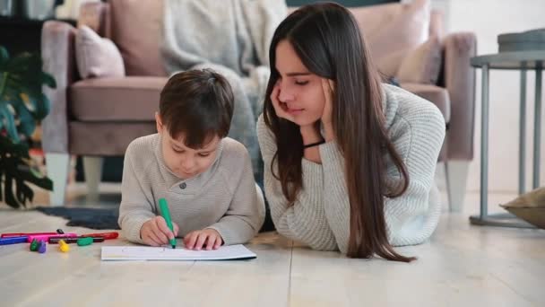 Bella giovane madre in un maglione caldo sdraiato sul pavimento con mio figlio che disegna con pennarelli su carta raffiguranti la sua famiglia. Il bambino impara a disegnare . — Video Stock