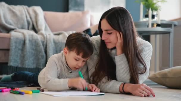 Hermosa madre joven en un suéter caliente acostado en el suelo con mi hijo dibujando con marcadores en papel que retratan a su familia. El niño aprende a dibujar . — Vídeo de stock