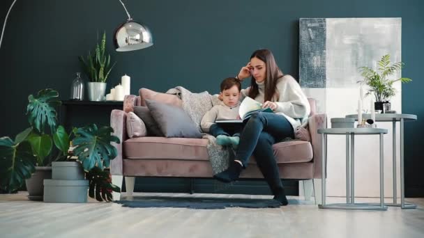Bela jovem mãe sentada no sofá em sua casa a sala de estar lê filho uma história no ouvido e aprender a ler menino . — Vídeo de Stock