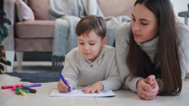 Oğlumla işaretleri ile ailesi tasvir kağıt üzerine çizim yerde yatarken bir sıcak kazaklı güzel genç anne. Çocuk çizmeyi öğrenir. Yakın çekim