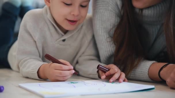 Beautiful young mother in a warm sweater lying on the floor with my son drawing with markers on paper portraying his family. The child learns to draw. Close-up — Stock Video