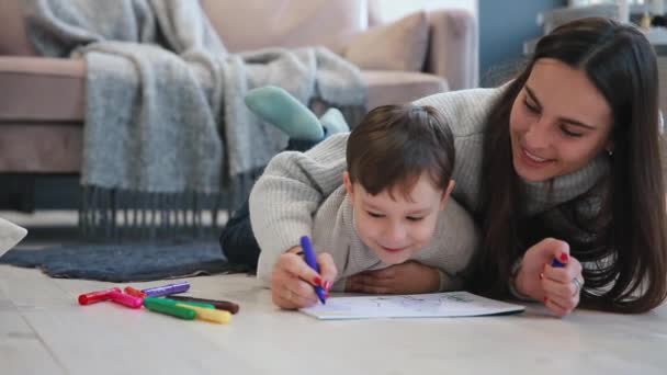 Krásná mladá matka v teplém svetru, ležící na podlaze s mým synem, kreslení se značkami na papíře ztvárnit svou rodinu. Dítě se učí kreslit. Detail — Stock video