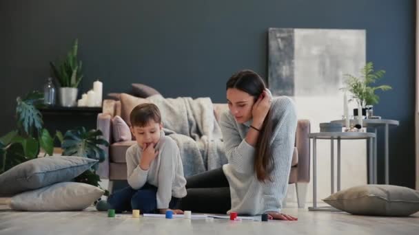 Mother with a child in the white interior of his house was opened the paint can sitting on the floor and dip your fingers in paint and draw on paper with your hands. Happy family. — Stock Video