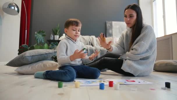 Mor med ett barn i vita insidan av hans hus öppnades måla burken sitter på golvet och doppa fingrarna i paint och rita på papper med händerna. Lycklig familj. — Stockvideo