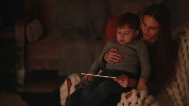Schöne Frau Mutter sitzt in einem Stuhl sein Haus mit Sohn auf dem Schoß beobachten Cartoons auf dem Touchscreen des Tablets. — Stockvideo