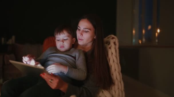 Vacker kvinna mor sitter i en stol sitt hem med son på hans knä titta på tecknade serier på pekskärmen av tabletten. — Stockvideo