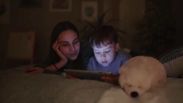 Glückliche Familienmutter und Sohn liegen auf dem Bett im Schlafzimmer seines Hauses und spielen Spiele auf dem Bildschirm des Tablets — Stockvideo