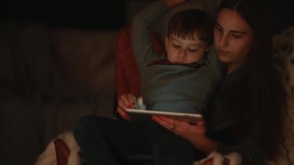 Όμορφη γυναίκα μητέρα κάθεται σε μια καρέκλα το σπίτι του, με γιο στην αγκαλιά του παρακολουθούν κινούμενα σχέδια στην οθόνη αφής του tablet. — Αρχείο Βίντεο