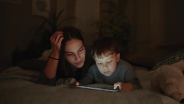 Счастливая семья мать и сын лежит на кровати в спальне своего дома, играя в игры экран планшета — стоковое видео